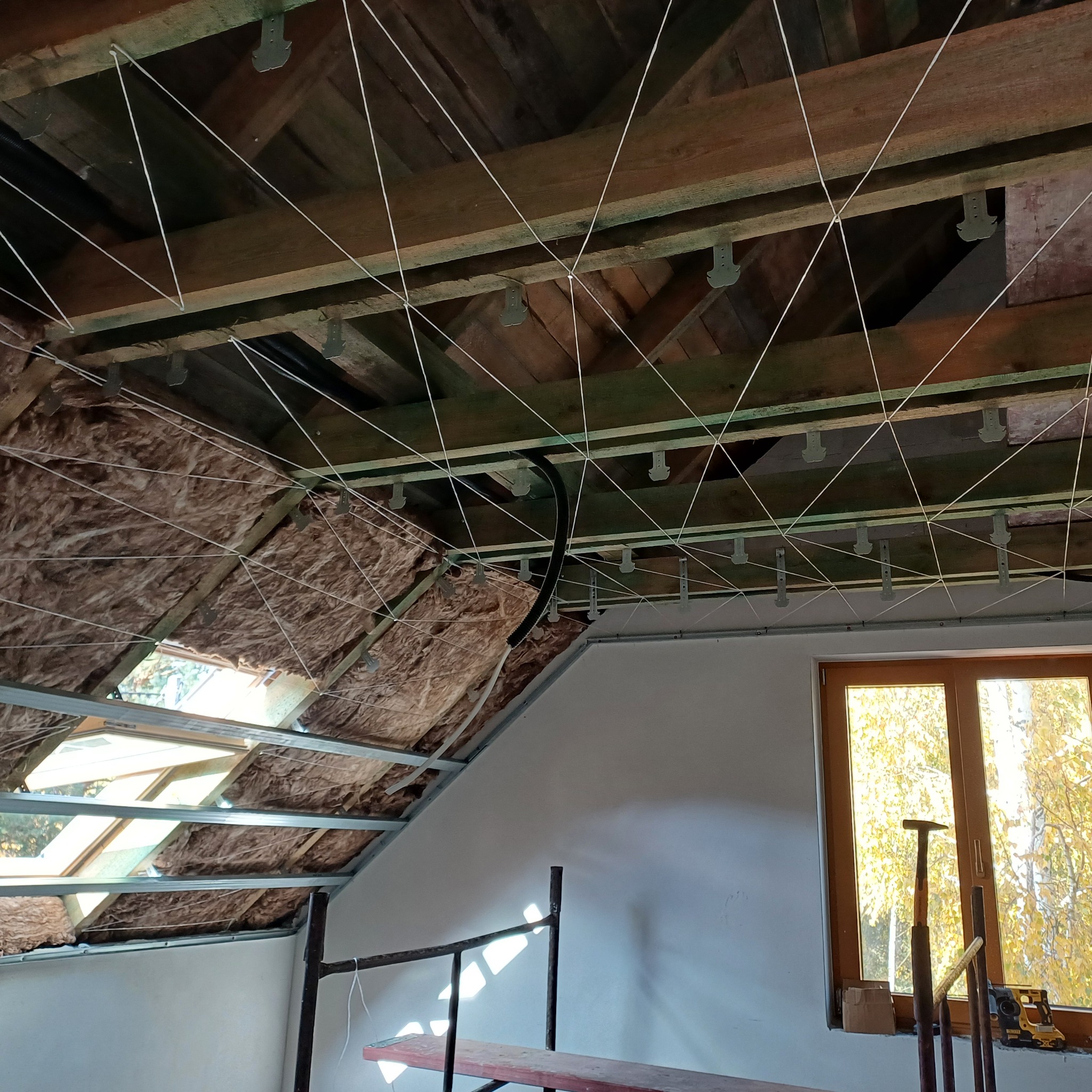 Ocieplenie poddasza wełną mineralną, widoczne drewniane krokwie, sznurki mocujące, okno dachowe i fragment rusztowania. Prace wykończeniowe w trakcie.