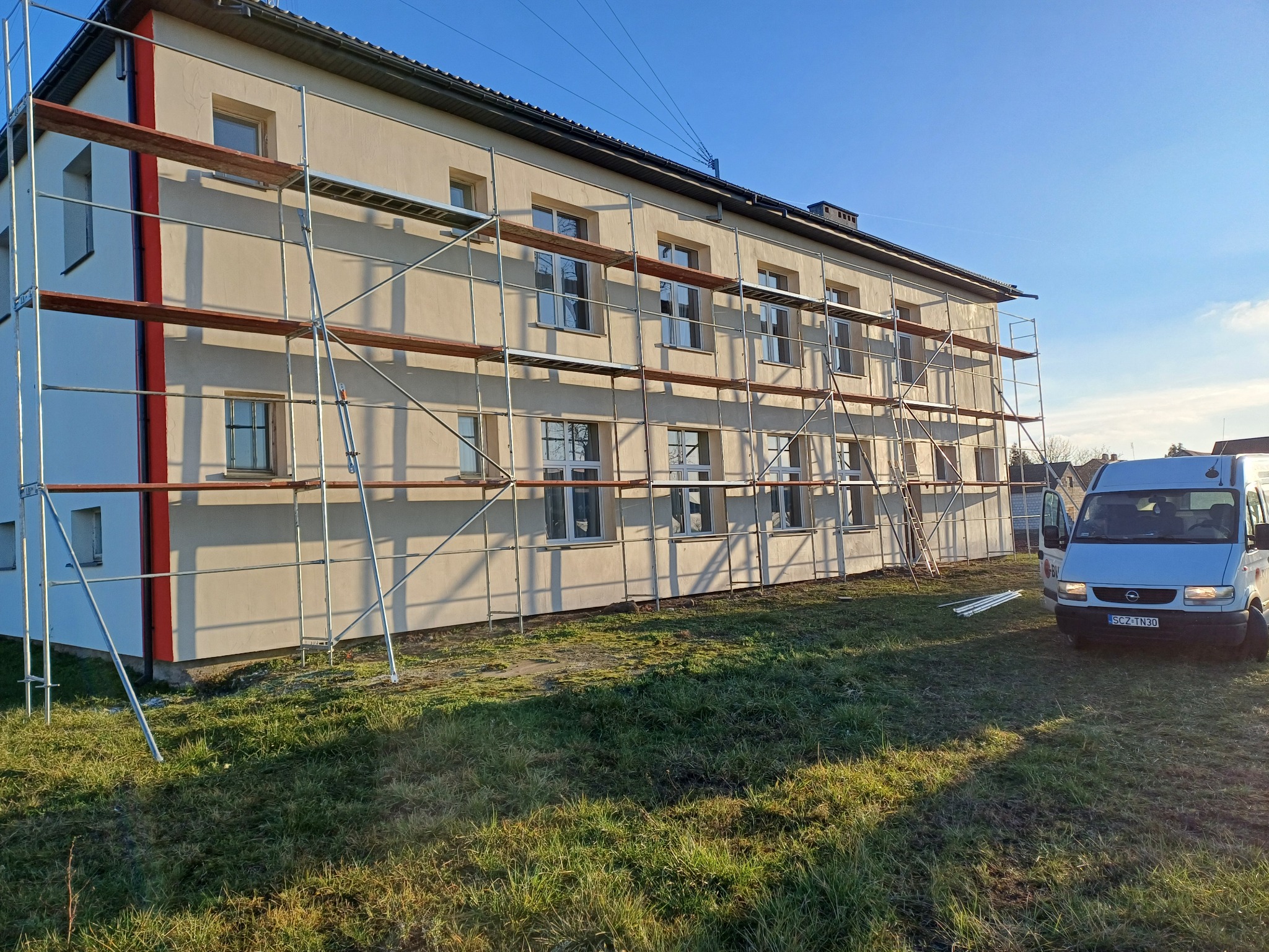 Budynek w trakcie prac elewacyjnych, otoczony rusztowaniem. Na pierwszym planie trawnik i zaparkowany biały bus. Słoneczny dzień.