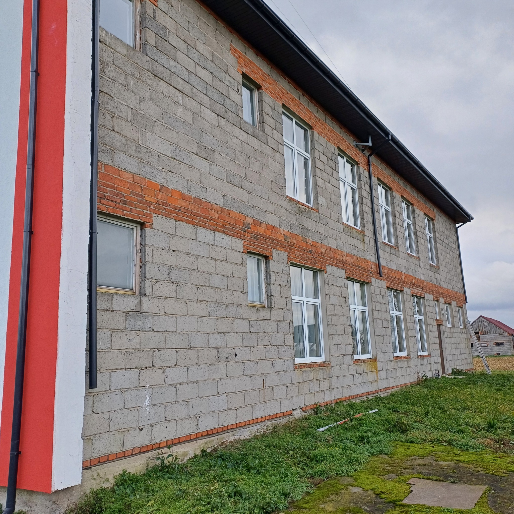 Surowa elewacja budynku z szarego bloczka, z pasami czerwonej cegły i białymi oknami. Fragment pomalowany w biało-czerwone pasy. Widoczne rynny i fragment dachu.