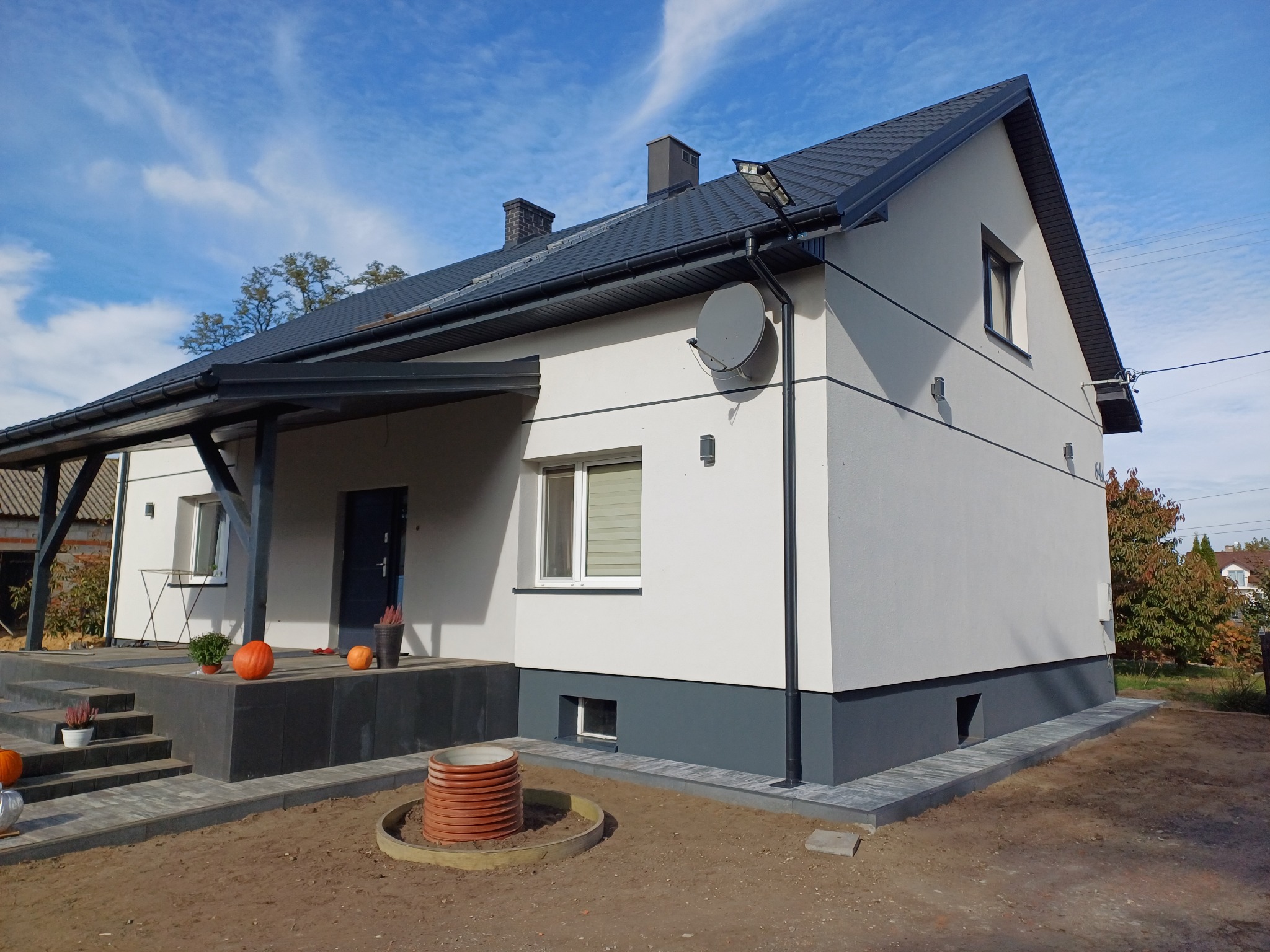 Nowoczesny dom z jasną elewacją i ciemnoszarą podmurówką, widoczne schody wejściowe i elementy dekoracyjne. Dach dwuspadowy z ciemnej dachówki. Błękitne niebo w tle.