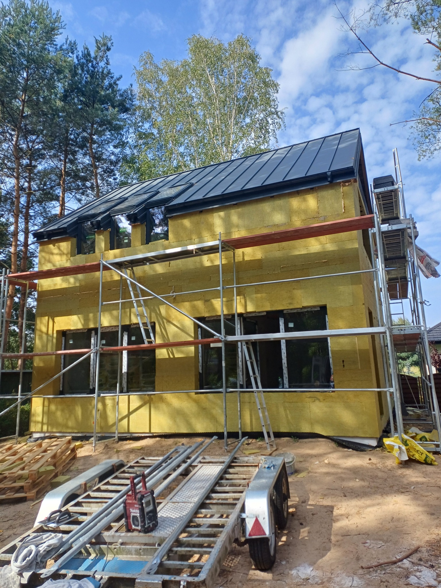 Dwukondygnacyjny dom w trakcie ocieplania, z widocznymi rusztowaniami i materiałem izolacyjnym w kolorze żółtym. Czarny dach i okna. Prace w toku.