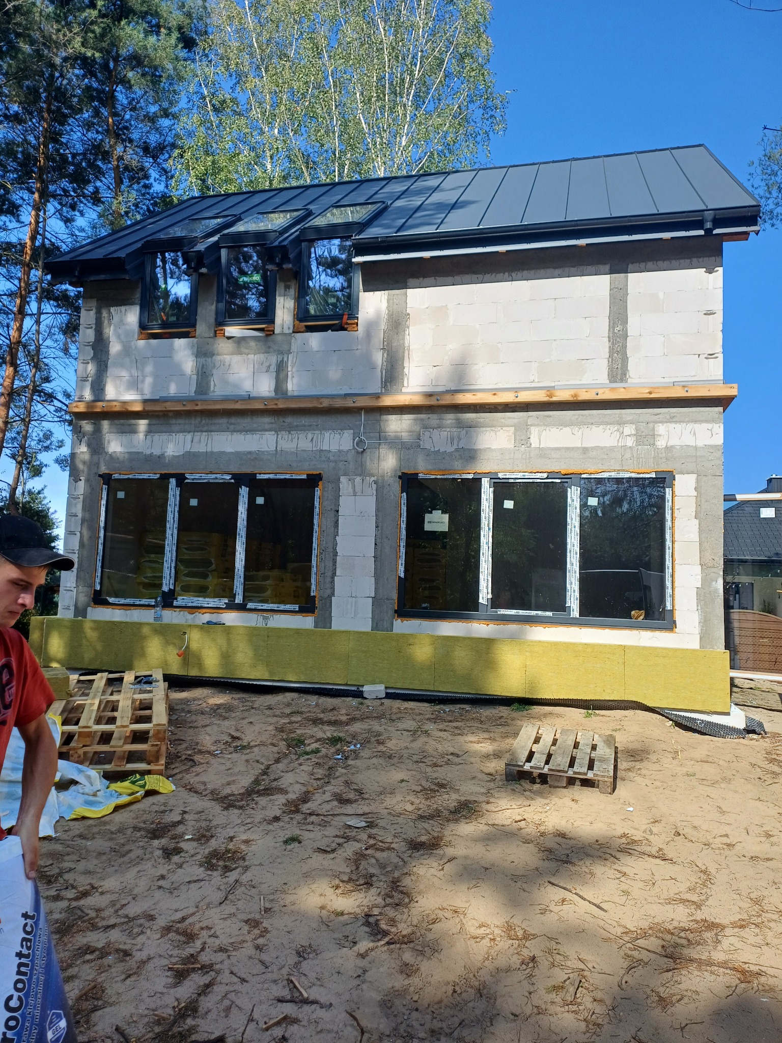 Surowy budynek z oknami i izolacją, widoczny fragment worka z zaprawą. Budowa domu w lesie, w trakcie prac elewacyjnych. Widoczne palety i piasek.