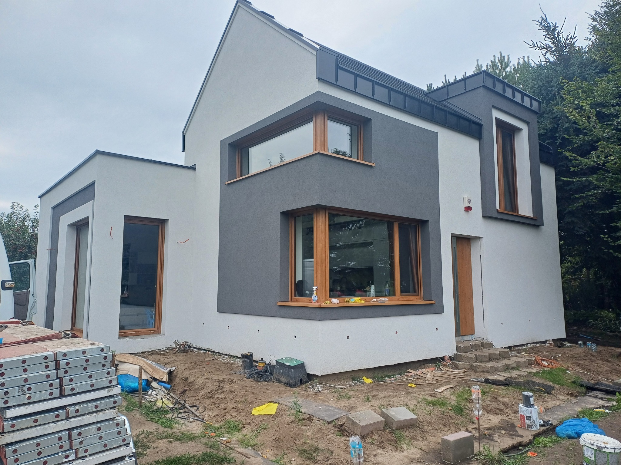 Nowoczesny dom z elewacją w odcieniach szarości i bieli, drewniane okna. Na pierwszym planie rusztowanie i teren po pracach budowlanych. Widok z zewnątrz.