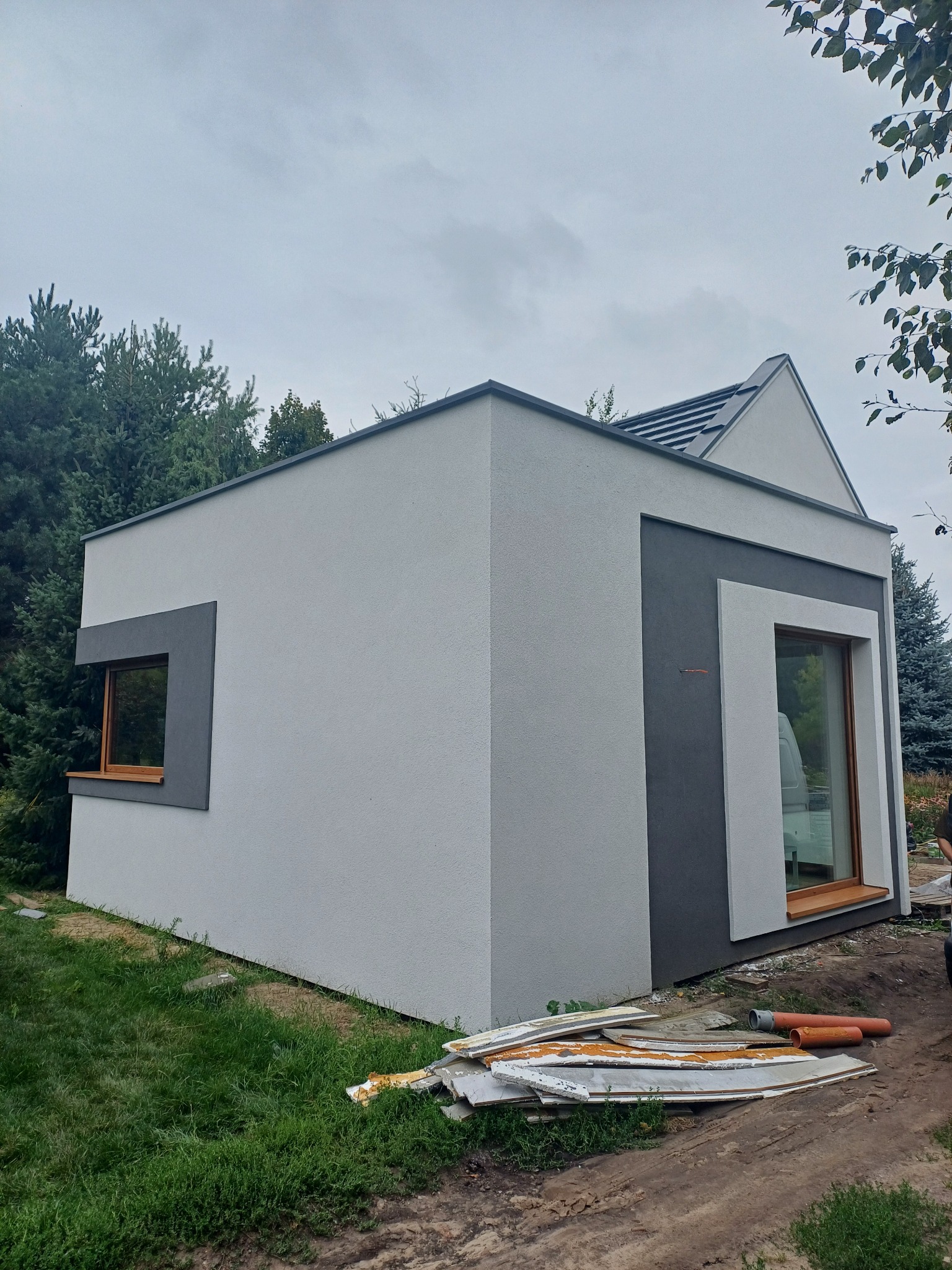 Nowoczesny, parterowy budynek z elewacją w odcieniach szarości i drewnianą stolarką okienną, otoczony zielenią. Widoczne ślady prac budowlanych.