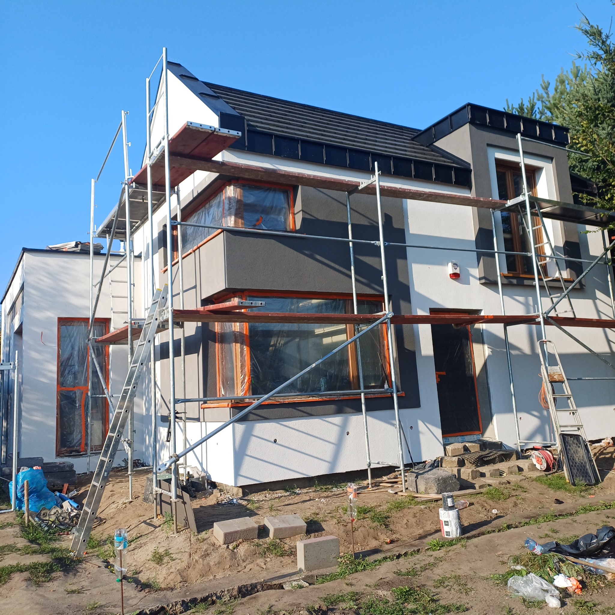 Dwukondygnacyjny dom w trakcie prac elewacyjnych. Widoczne rusztowania, zabezpieczone okna folią, częściowo pomalowana fasada w odcieniach bieli i szarości.