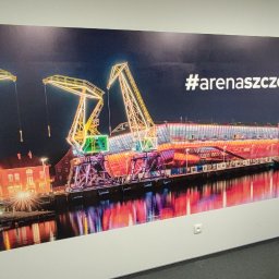DrukUp - Duży wydruk fotograficzny na ścianie przedstawia nocny widok iluminowanych dźwigów portowych i budynku, z odbiciami w wodzie i napisem #arenaszczecin, obok fragment czarnej sofy.
