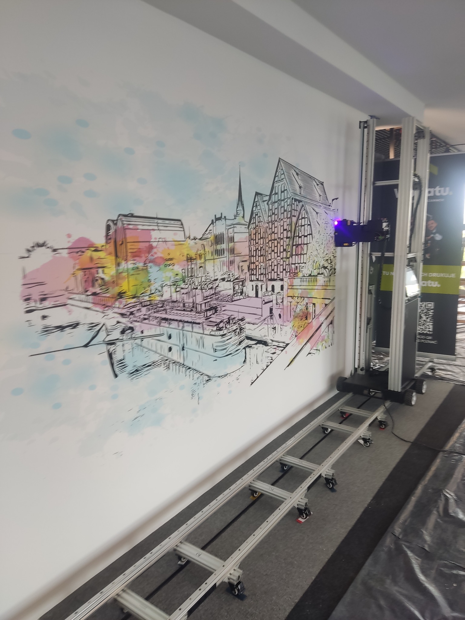 Ściana z nadrukowanym kolorowym szkicem panoramy miasta z rzeką, wykonanym za pomocą specjalistycznej drukarki pionowej poruszającej się na szynach.