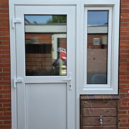 KMSI GROUP SPÓŁKA Z OGRANICZONĄ ODPOWIEDZIALNOŚCIĄ - Białe drzwi PCV z bocznym oknem zamontowane w murze z czerwonej cegły, z widocznym odbiciem osoby w szybie i drewnianą skrzynką z kranem u dołu.