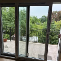 Montaż okna balkonowego z białymi ramami Aluplast. Widok na zieleń z balkonu. Pracownik uszczelnia pianką montażową. Jasne wnętrze.