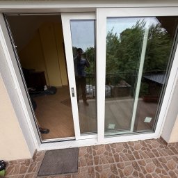 KMSI GROUP SPÓŁKA Z OGRANICZONĄ ODPOWIEDZIALNOŚCIĄ - Białe przesuwne okno balkonowe z widokiem na zewnątrz. Widoczny fragment balkonu z roślinnością i osoba robiąca zdjęcie w odbiciu szyby.