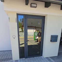 Szare drzwi zewnętrzne z szybą, zamontowane w wejściu do budynku. Widoczna skrzynka na listy i kostka brukowa.