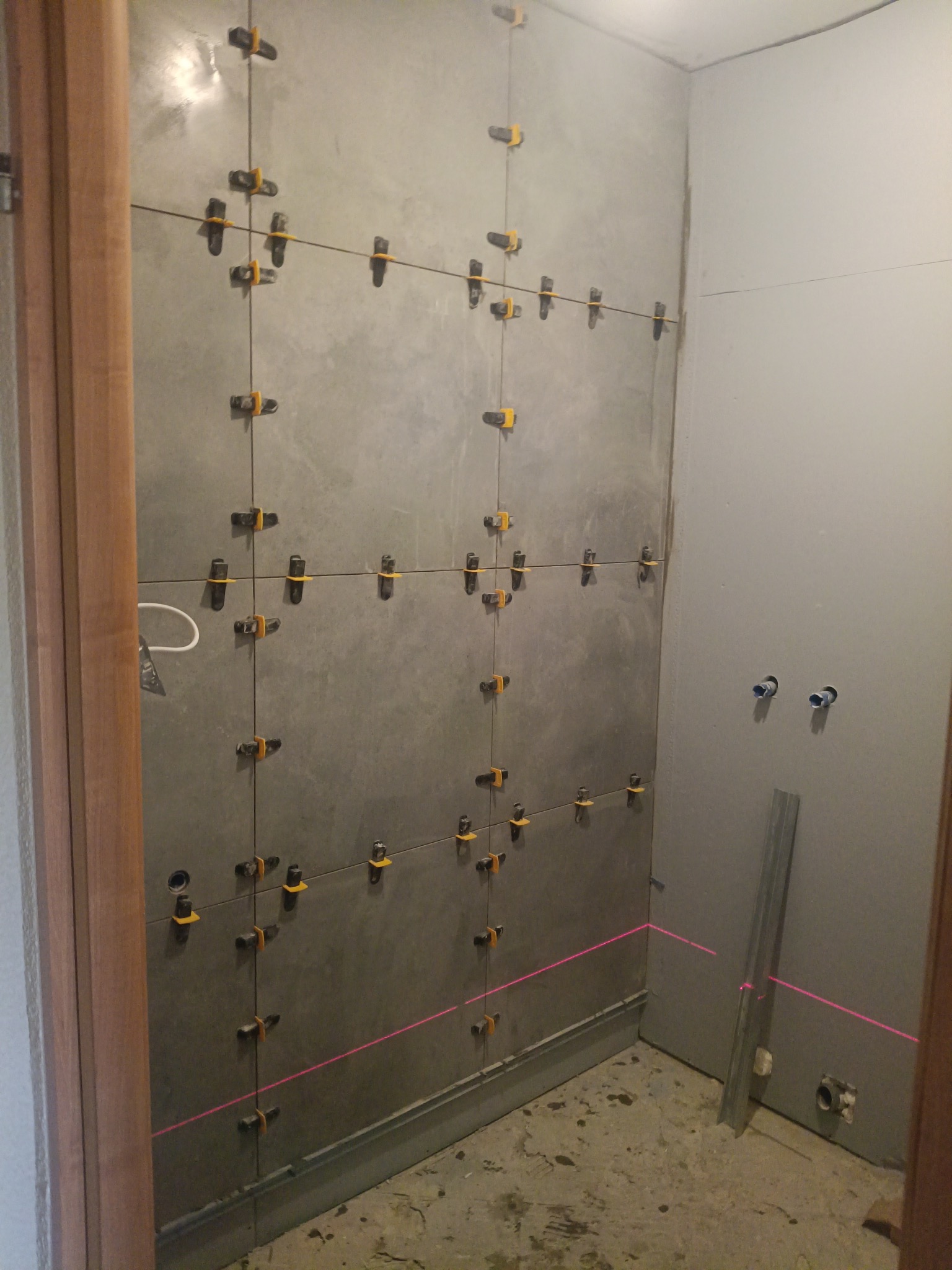 Ściana w trakcie układania szarych płytek z użyciem systemu poziomowania, widoczna linia lasera wyznaczająca poziom, surowy stan wykończenia łazienki.