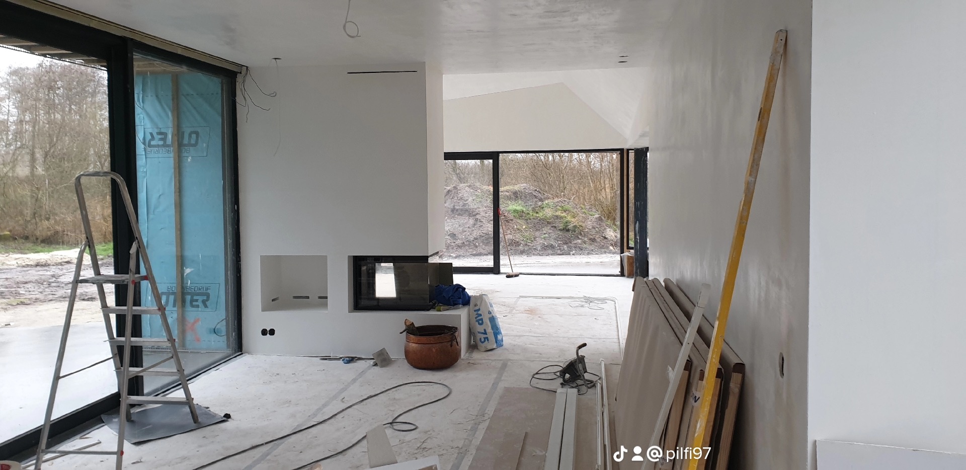 Wnętrze w trakcie remontu z widocznymi oknami z czarnymi ramami, przygotowaną ścianą pod malowanie, schodami, poziomnicą, workiem gipsu MP 75 oraz elementami zabudowy kominka.