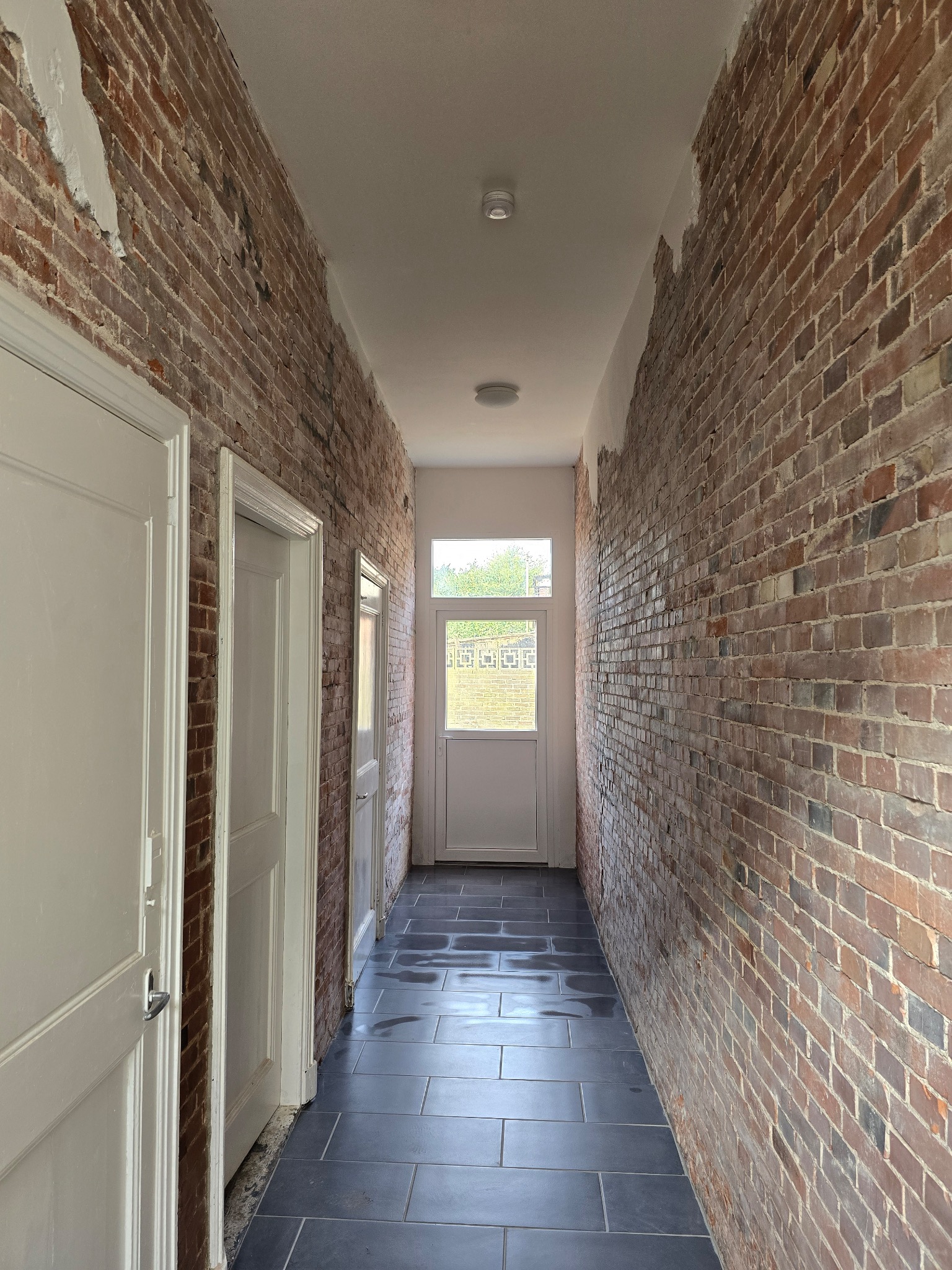 Wąski korytarz z ceglanymi ścianami w trakcie remontu, widoczne ślady po usuniętym tynku, nowe ciemne płytki na podłodze, białe drzwi na końcu korytarza wpuszczające światło dzienne.