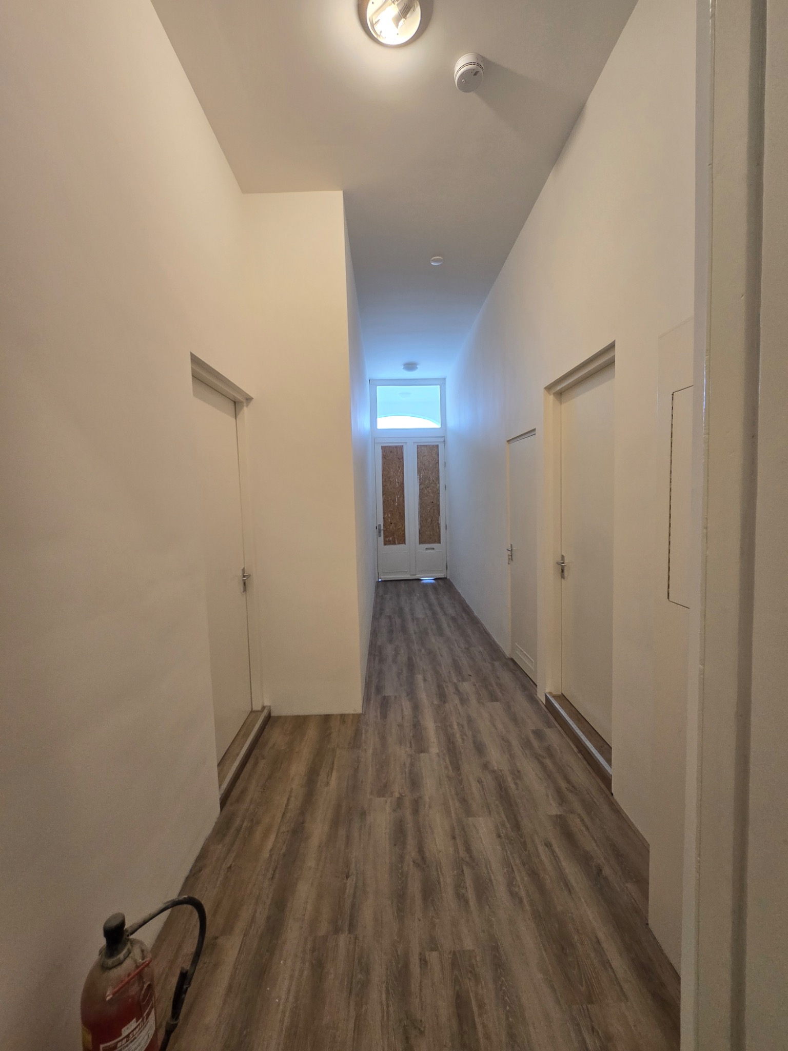 Długi, jasny korytarz z panelami podłogowymi imitującymi drewno, białymi ścianami i drzwiami, z widocznym gaśnicą w dolnym rogu kadru.