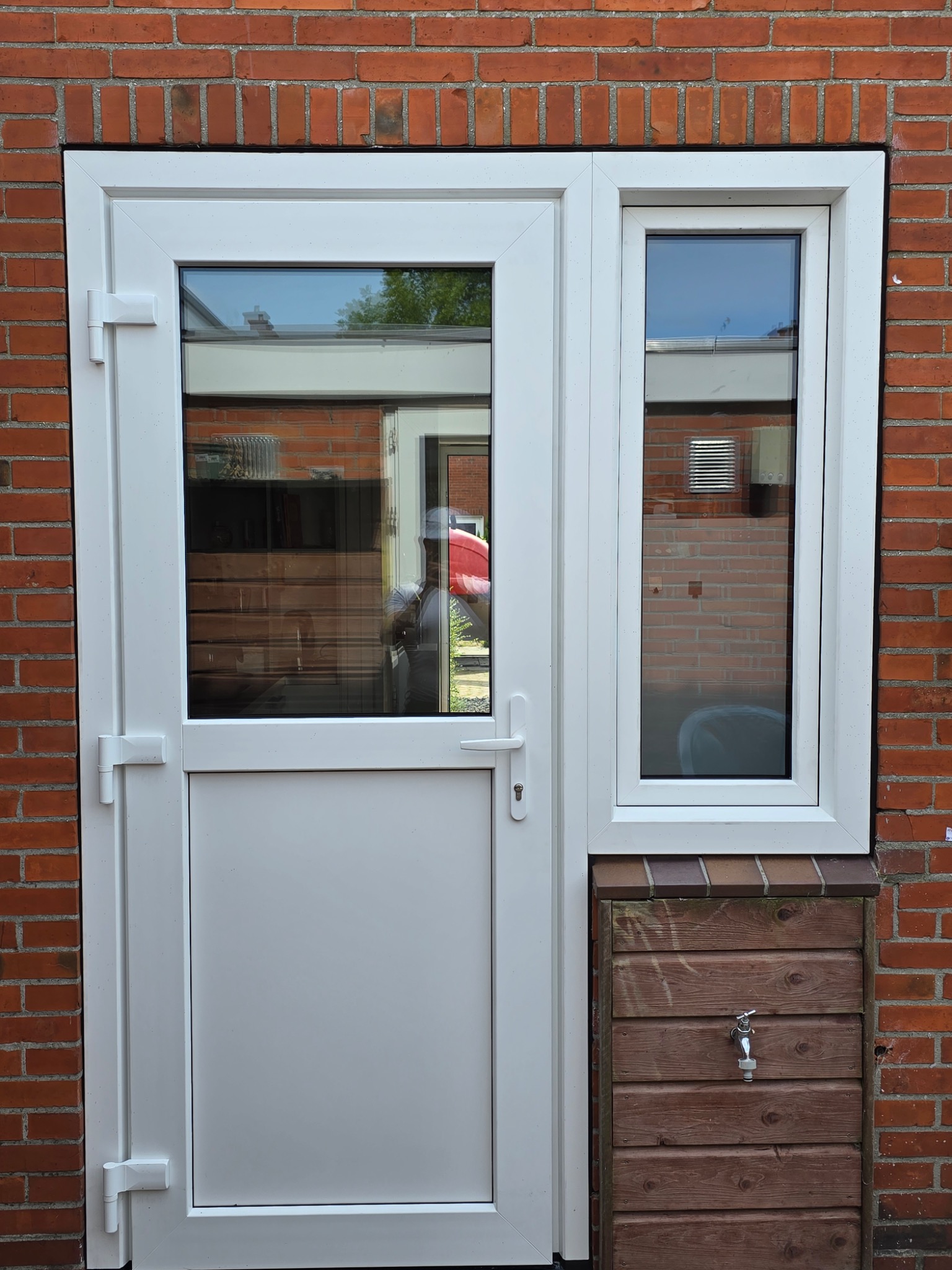Białe drzwi PCV z bocznym oknem zamontowane w murze z czerwonej cegły, z widocznym odbiciem osoby w szybie i drewnianą skrzynką z kranem u dołu.