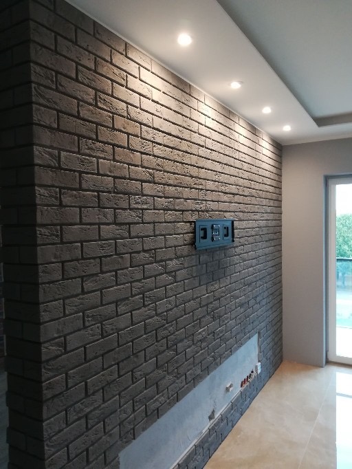 Ściana z ciemnej cegły dekoracyjnej z ukrytymi przewodami i uchwytem na telewizor, oświetlona punktowymi lampami sufitowymi.