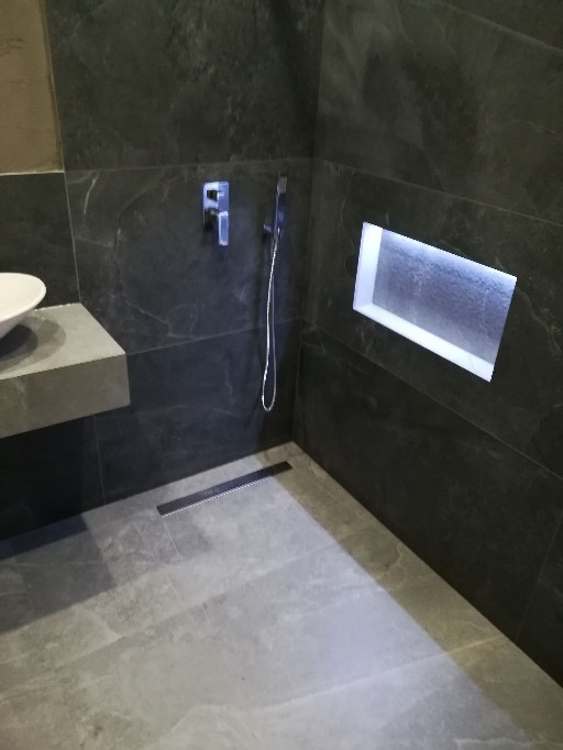 Nowoczesna kabina prysznicowa z ciemnoszarą, marmurkową okładziną ścienną, minimalistyczną armaturą i odpływem liniowym w posadzce z szarych płytek.