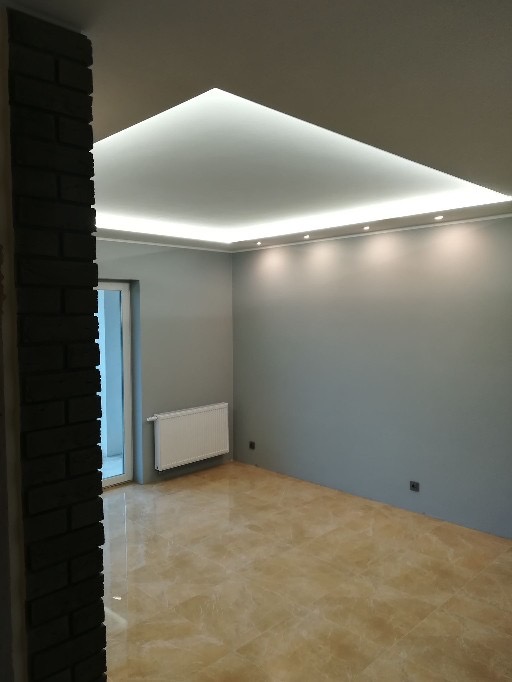 Wnętrze pokoju z marmurową podłogą, szarą ścianą, grzejnikiem i podwieszanym sufitem z oświetleniem LED, widoczne fragmenty ściany obłożonej cegłą.