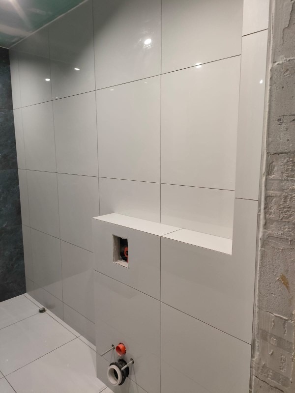 Białe, błyszczące płytki ceramiczne ułożone na ścianie łazienki z wnęką i przygotowanymi przyłączami hydraulicznymi, widoczne ślady prac wykończeniowych na sąsiedniej ścianie.