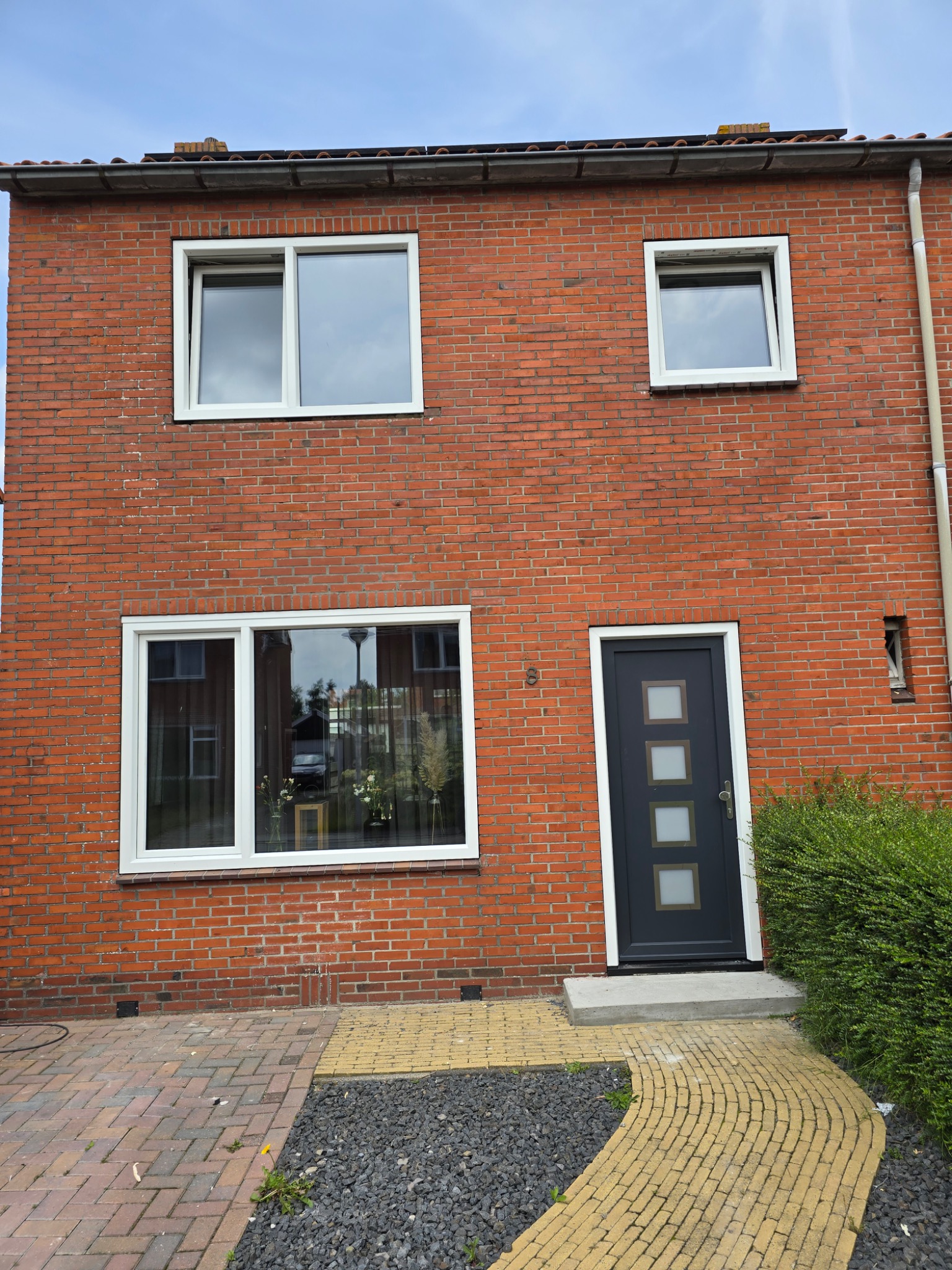 Dwukondygnacyjny dom z czerwonej cegły z trzema białymi oknami i ciemnoszarymi drzwiami z trzema kwadratowymi przeszkleniami, otoczony kostką brukową i krzewami.