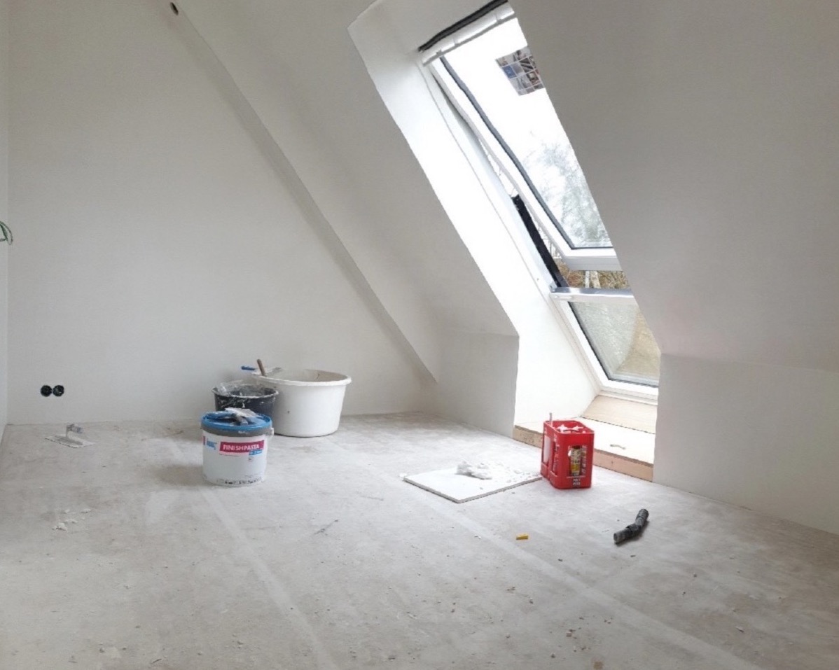 Pokój na poddaszu w trakcie prac wykończeniowych; widoczne wiadra z farbą, szpachla, okno dachowe i czerwona skrzynka na napoje.