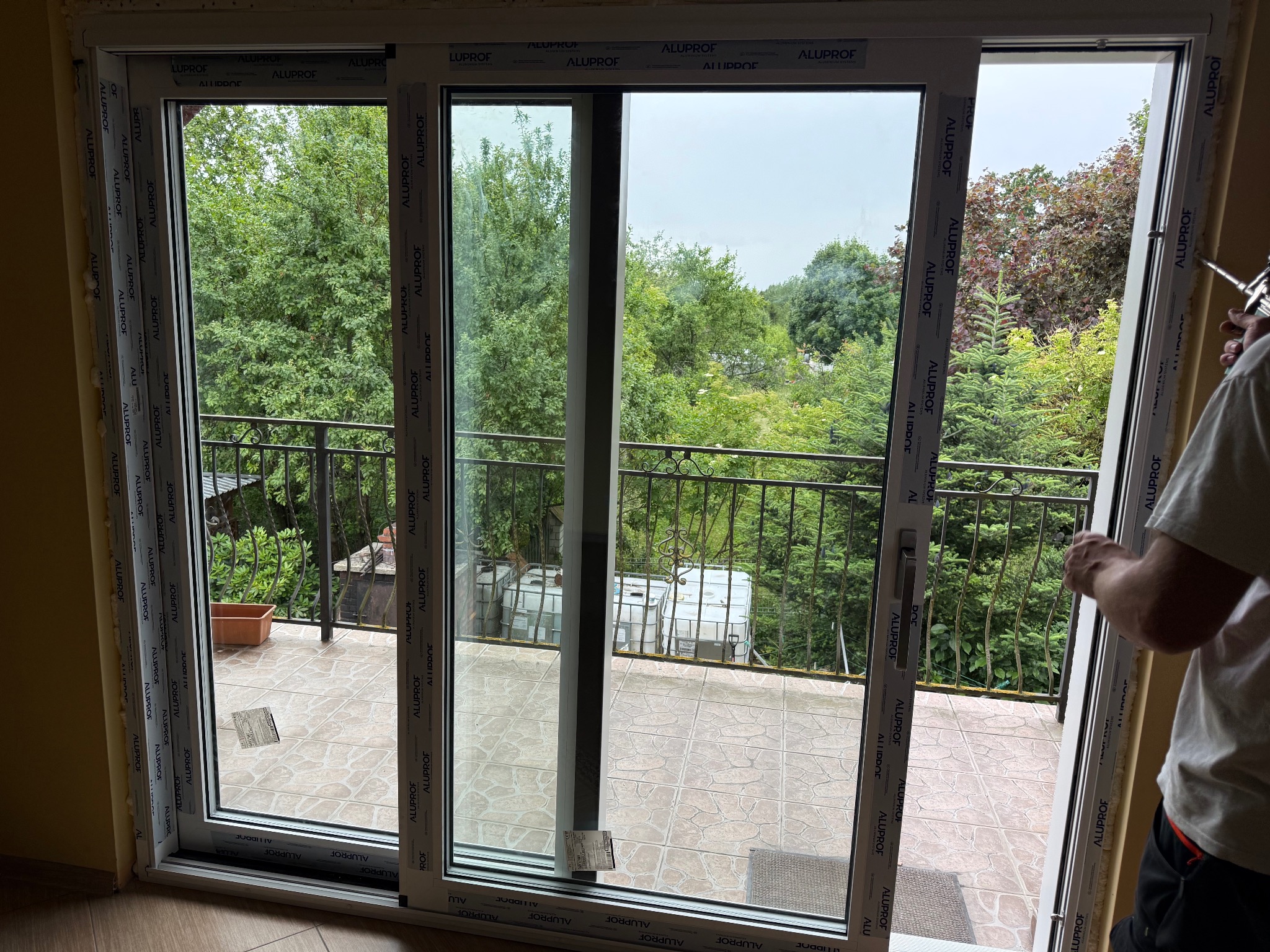 Montaż okna balkonowego z białymi ramami Aluplast. Widok na zieleń z balkonu. Pracownik uszczelnia pianką montażową. Jasne wnętrze.