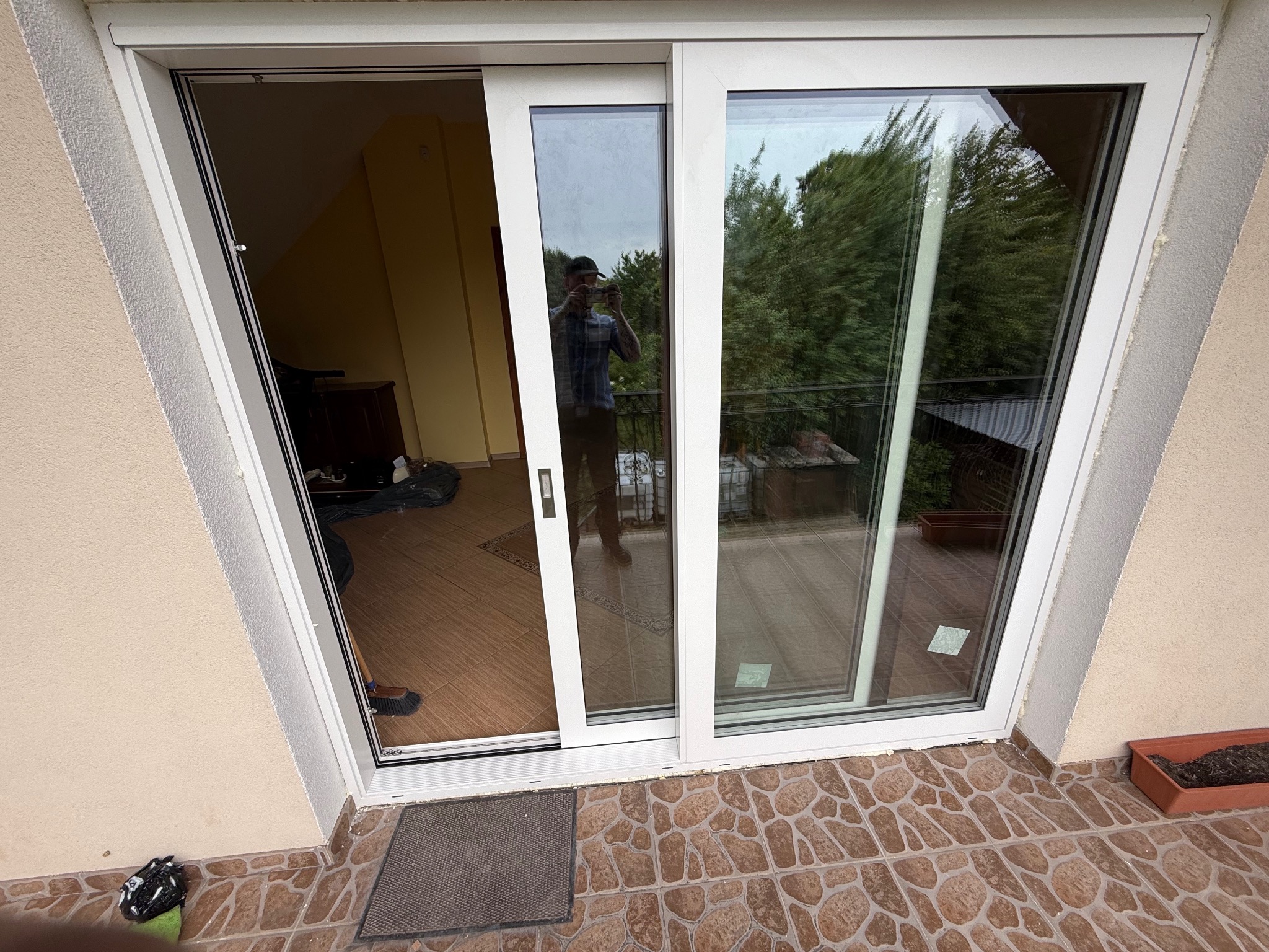 Białe przesuwne okno balkonowe z widokiem na zewnątrz. Widoczny fragment balkonu z roślinnością i osoba robiąca zdjęcie w odbiciu szyby.