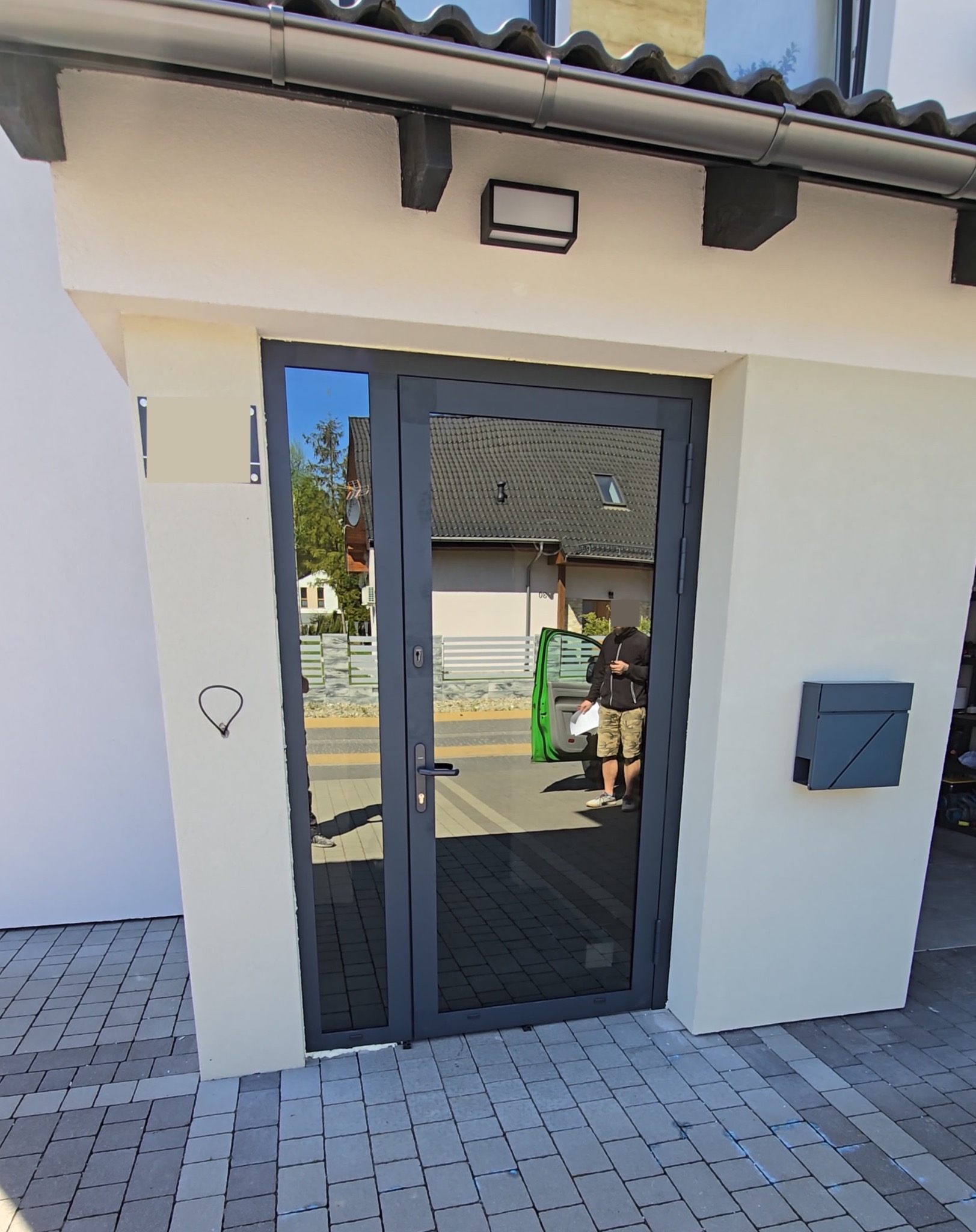 Szare drzwi zewnętrzne z szybą, zamontowane w wejściu do budynku. Widoczna skrzynka na listy i kostka brukowa.