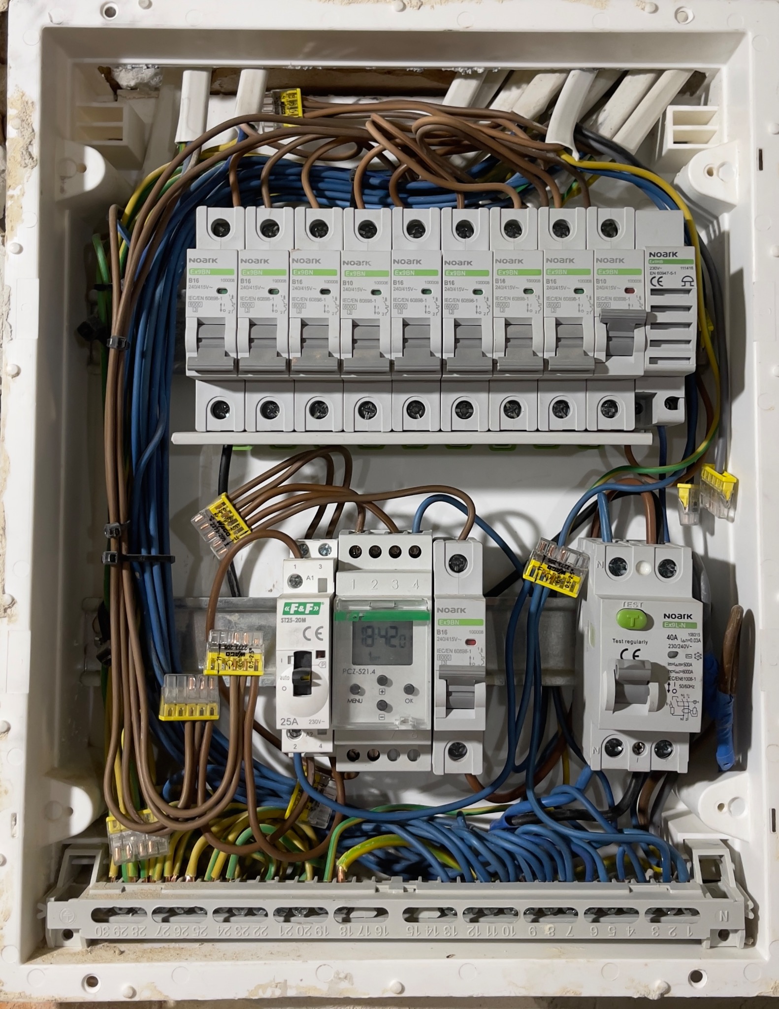 Rozdzielnia elektryczna w białej obudowie z widocznymi bezpiecznikami, przewodami i elementami sterującymi. Widoczne złącza WAGO i timer.