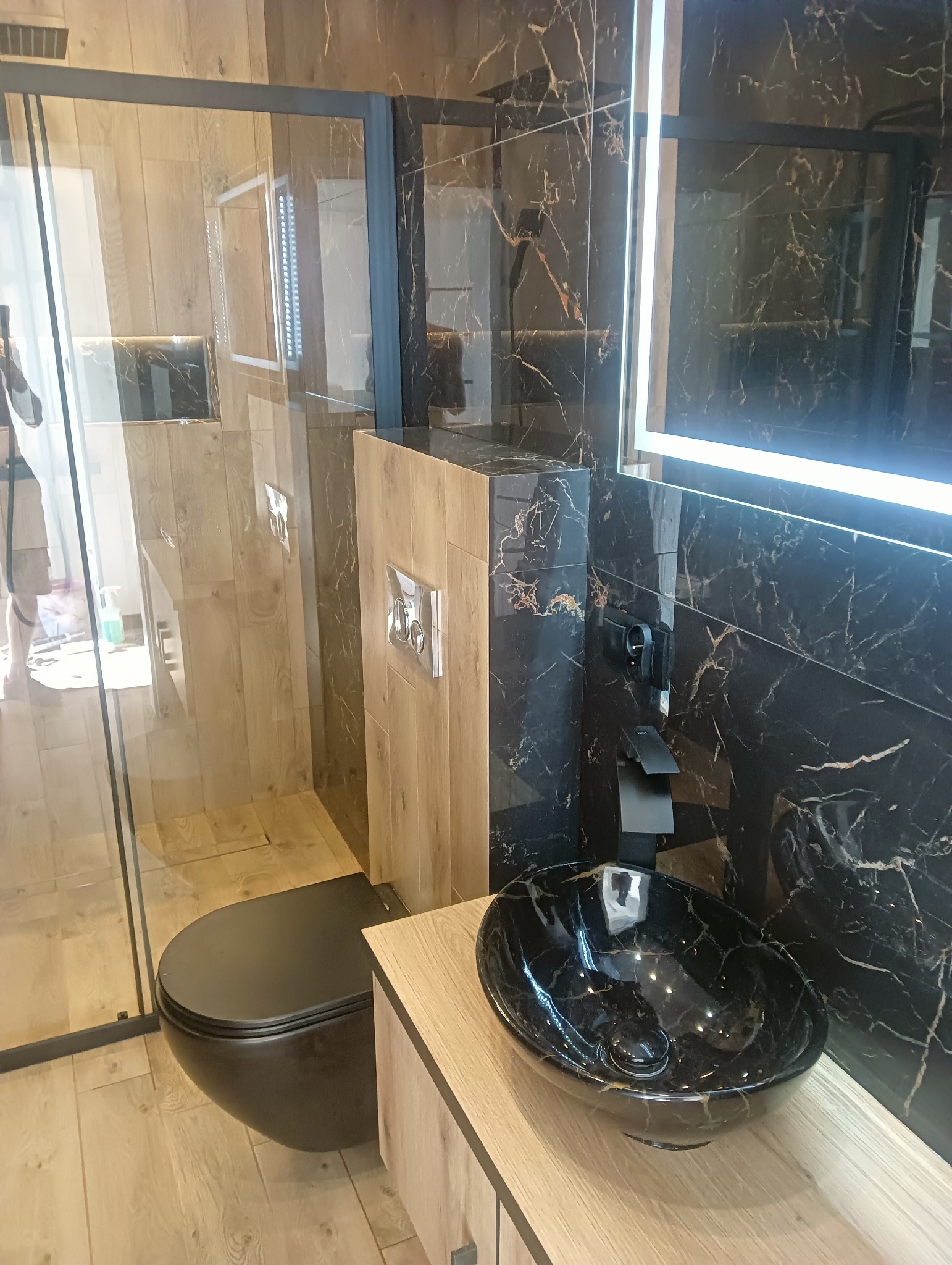 Nowoczesna łazienka z czarną armaturą i umywalką w marmurkowy wzór, drewnopodobne płytki na podłodze i ścianach, oraz podświetlane lustro LED.