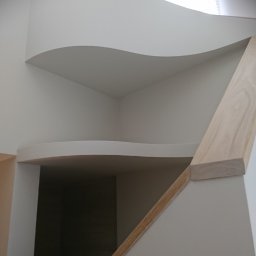 KUZ-BUD - Nietypowa, falista zabudowa z płyt karton-gips w jasnym wnętrzu, z widoczną drewnianą listwą i kwiatami w tle.