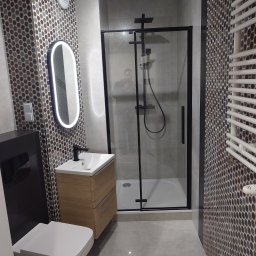 KUZ-BUD - Nowoczesna łazienka z kabiną prysznicową w czarnej ramie, umywalką z szafką w kolorze drewna, podświetlanym lustrem owalnym i mozaiką na ścianach.