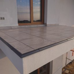 KUZ-BUD - Wykończony taras z szarych płytek ceramicznych na zewnątrz budynku, widoczne okno z drewnianą ramą i fragment rusztowania.