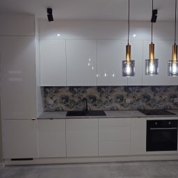 KUZ-BUD - Nowoczesna kuchnia z białymi, lakierowanymi frontami, szarym blatem i dekoracyjną tapetą w kwiaty nad blatem, oświetlona trzema wiszącymi lampami z elementami złota i przezroczystego szkła.