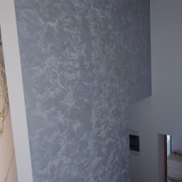 KUZ-BUD - Ściana wewnątrz budynku pokryta dekoracyjną szarą farbą z efektem pociągnięć pędzla, widoczne przewody elektryczne w rogu pomieszczenia.