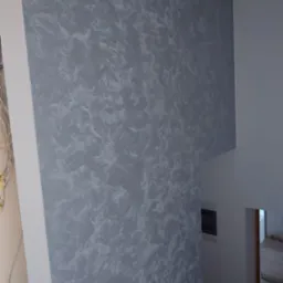 Ściana wewnątrz budynku pokryta dekoracyjną szarą farbą z efektem pociągnięć pędzla, widoczne przewody elektryczne w rogu pomieszczenia.