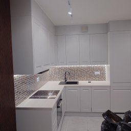 KUZ-BUD - Jasnoszara kuchnia z mozaiką na ścianie, czarnym zlewem i płytą indukcyjną, widoczne listwy dekoracyjne na ścianie i worki na śmieci na podłodze.