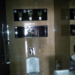 KUZ-BUD - Wykończona łazienka z beżowymi płytkami, podwieszaną toaletą, umywalką i dekoracyjnymi wnękami z roślinami i szklaną kulą.