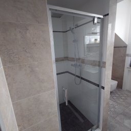 KUZ-BUD - Nowoczesna kabina prysznicowa z przesuwnymi drzwiami i deszczownicą, wykończona płytkami w odcieniach beżu i szarości, z widocznym odpływem liniowym w podłodze.