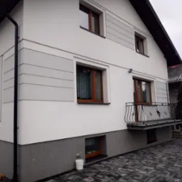 Dwupiętrowy dom jednorodzinny po świeżym ociepleniu elewacji w Radomiu, z widoczną strukturą tynku i dekoracyjnymi szarymi pasami, balkon z kutą balustradą, kostka brukowa na podjeździe i białe...