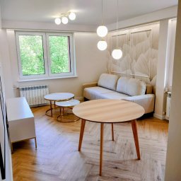 KUZ-BUD - Jasne wnętrze po wykończeniu: parkiet w jodełkę, okrągły stół, sofa, stoliki kawowe, dekoracyjna ściana z motywem liści, nowoczesne lampy.