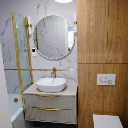 KUZ-BUD - Wykończona łazienka w bieli i drewnie: umywalka na szafce z frezowanymi frontami, okrągłe lustro, kabina prysznicowa w złotych ramach, WC z drewnianą zabudową.