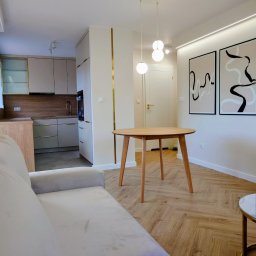 KUZ-BUD - Jasne wnętrze po remoncie: parkiet w jodełkę, okrągły stół z drewna, beżowa sofa i minimalistyczne grafiki na ścianie. Kuchnia w tle, z zabudową w odcieniach beżu i drewna.