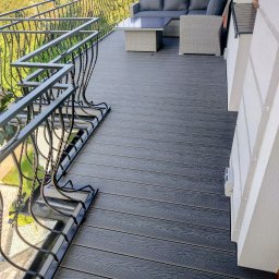 KUZ-BUD - Stylowy balkon z szarą podłogą z desek kompozytowych, czarną, kutą balustradą i kompletem wypoczynkowym w tle. Widok na zieleń i domy.