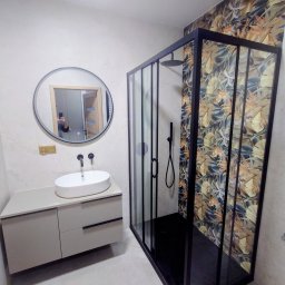 KUZ-BUD - Nowoczesna łazienka z kabiną prysznicową w czarnej ramie i wzorzystą tapetą, okrągłe lustro nad umywalką na szafce w odcieniach szarości, minimalistyczne baterie.