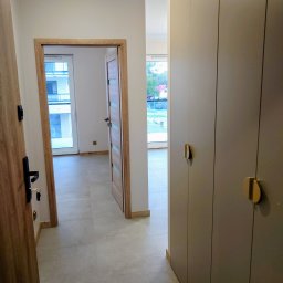 KUZ-BUD - Nowoczesny korytarz z beżową podłogą, drewnianymi drzwiami i dużą szafą w zabudowie z złotymi uchwytami. Widok na okno i dalszą część mieszkania.