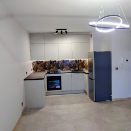 KUZ-BUD - Nowoczesna kuchnia w odcieniach szarości z dekoracyjnym panelem nad blatem. Oświetlenie punktowe i designerska lampa wisząca. Widok na fragment salonu.