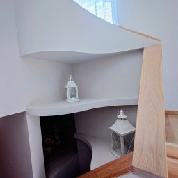 KUZ-BUD - Białe, zaokrąglone półki wnękowe z dekoracyjnymi lampionami, drewniane elementy schodów, minimalistyczny design, jasne wnętrze z oknem w tle.