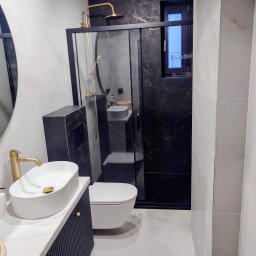KUZ-BUD - Nowoczesna łazienka z kabiną typu walk-in, czarnymi płytkami imitującymi marmur i złotą armaturą. Umywalka nablatowa na szafce z ryflowanym frontem.